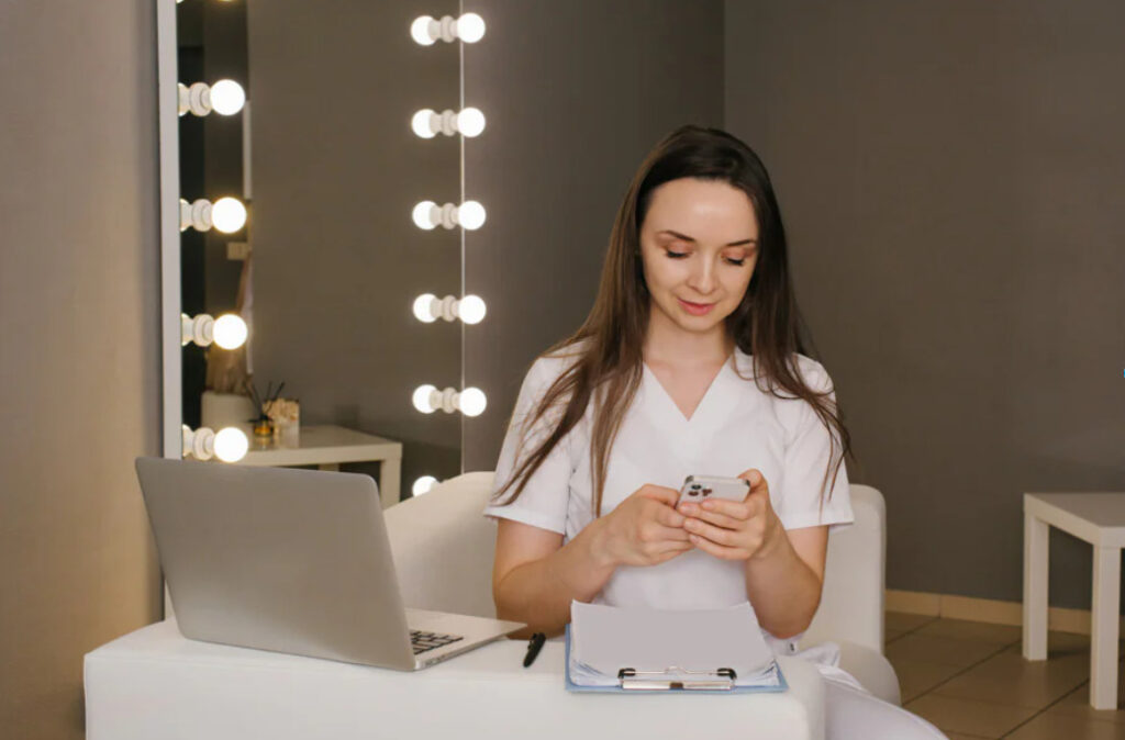 Mulher com celular nas mãos e notebook sobre a mesa fazendo referência a onde aprender a tratar Eflúvio Anágeno.