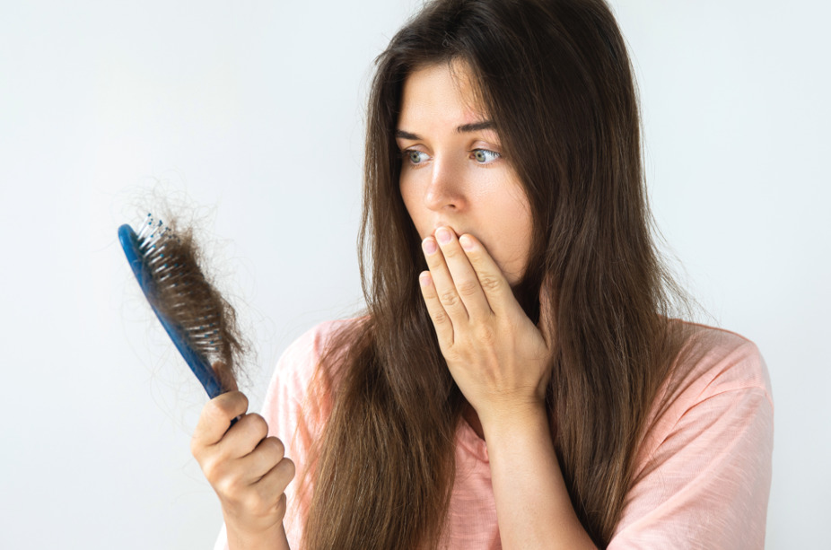 Mulher olhando preocupada para os fios de cabelo na escova, fazendo referência a causas da queda de cabelo.
