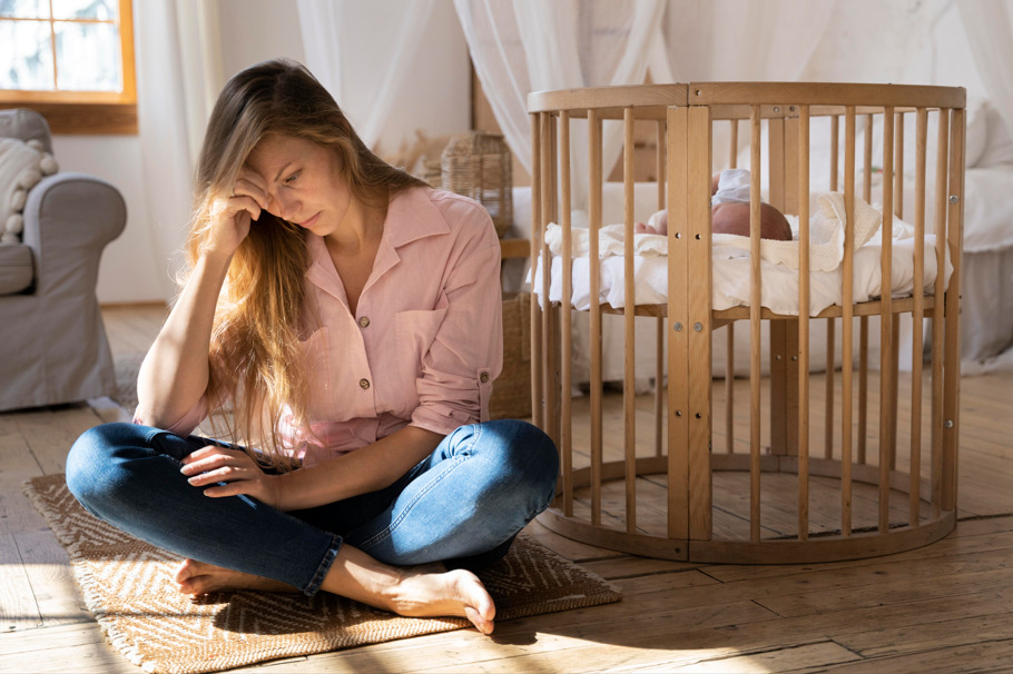 Mulher sentada perto do berço preocupada com queda de cabelo pós-parto.